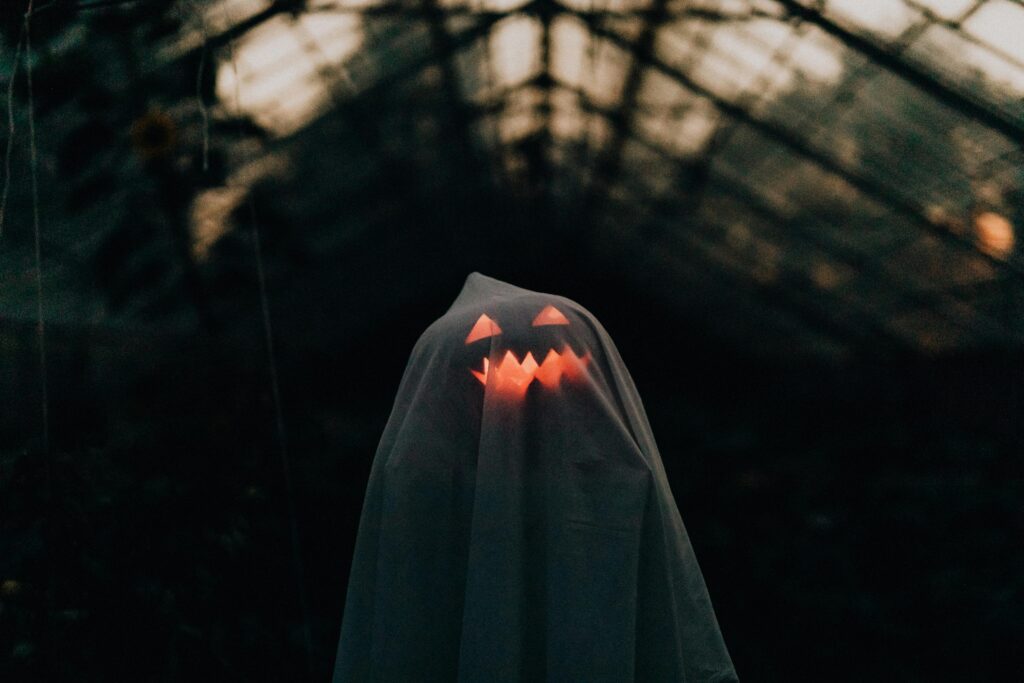 A ghostly figure with a glowing pumpkin face under a sheet in an eerie, dark setting. Perfect Halloween imagery.